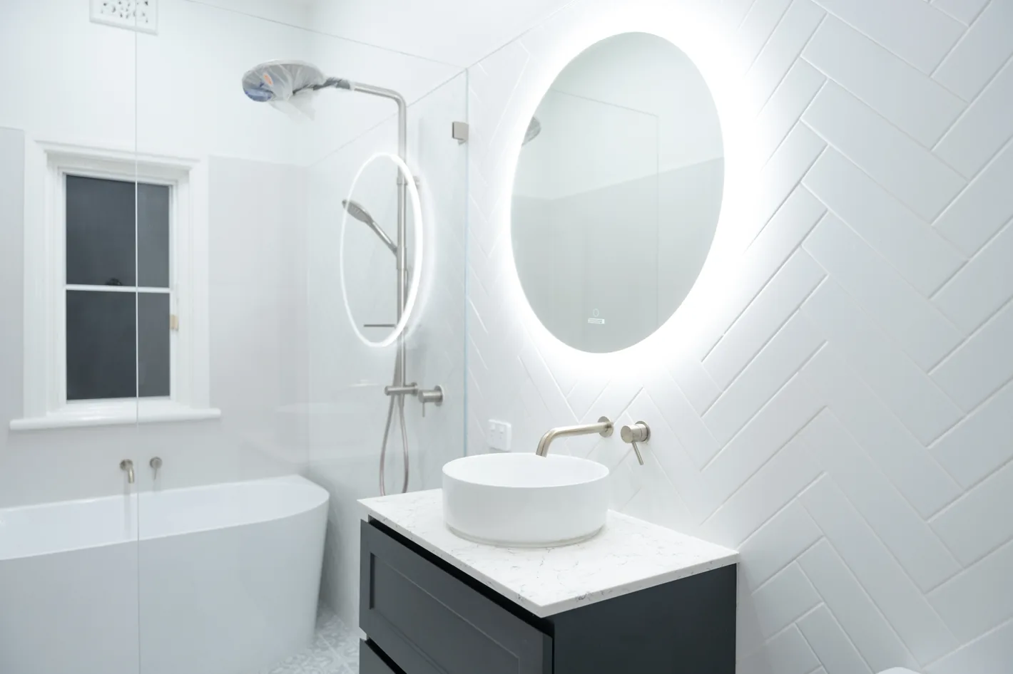 Completed bathroom renovation with herringbone wall tiling, round backlit mirror, freestanding basin and bath