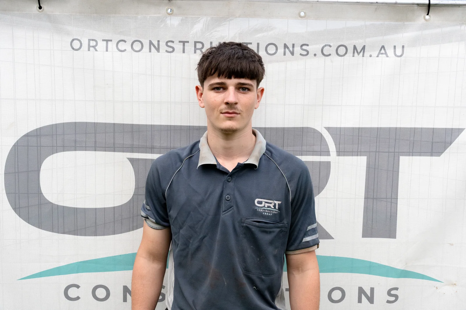 Alyster, apprentice carpenter at ORT Constructions, in front of an ORT Constructions site banner
